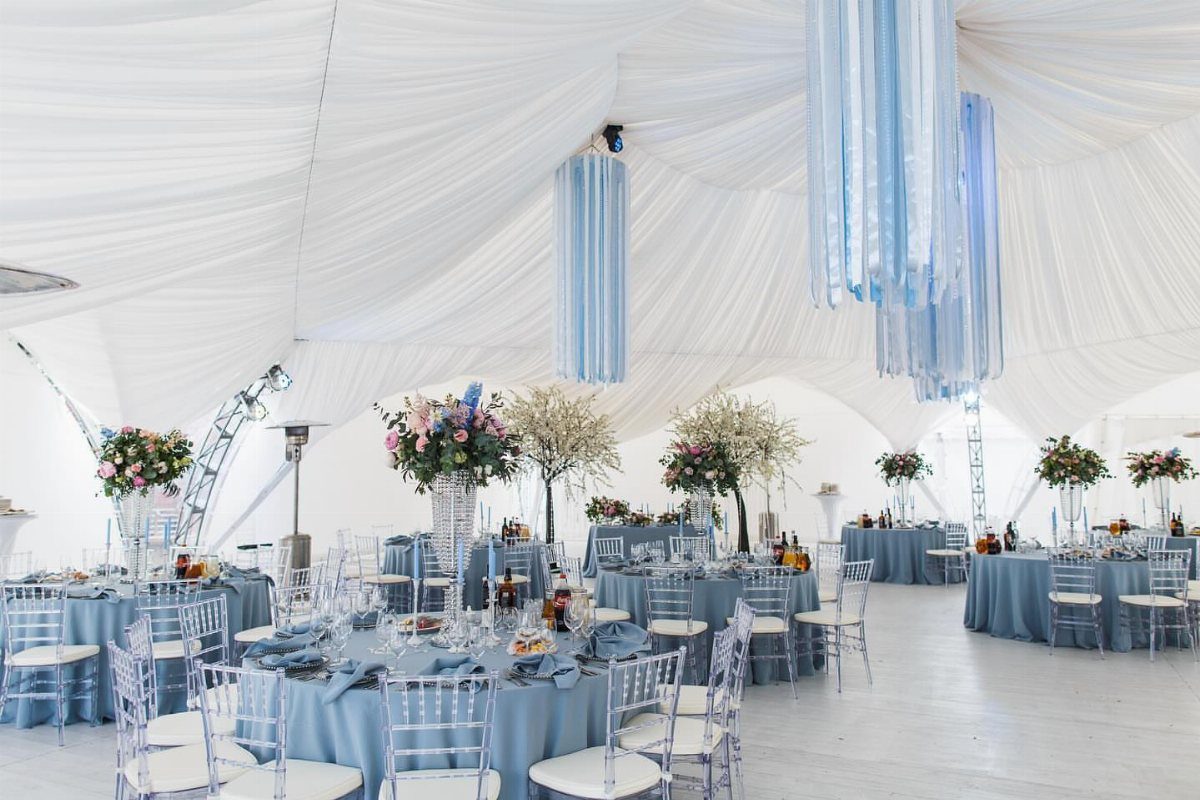 Light blue table linens and blue hanging decorations inside a white wedding tent.