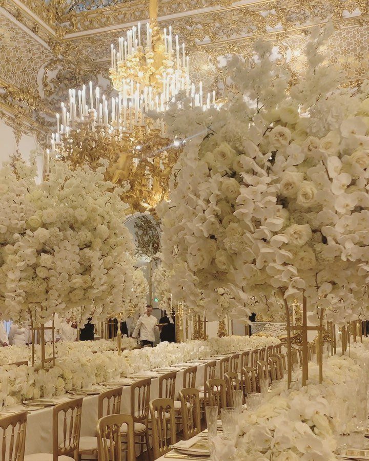 Lavish ballroom with gold decor, crystal chandeliers, and massive white floral centerpieces.