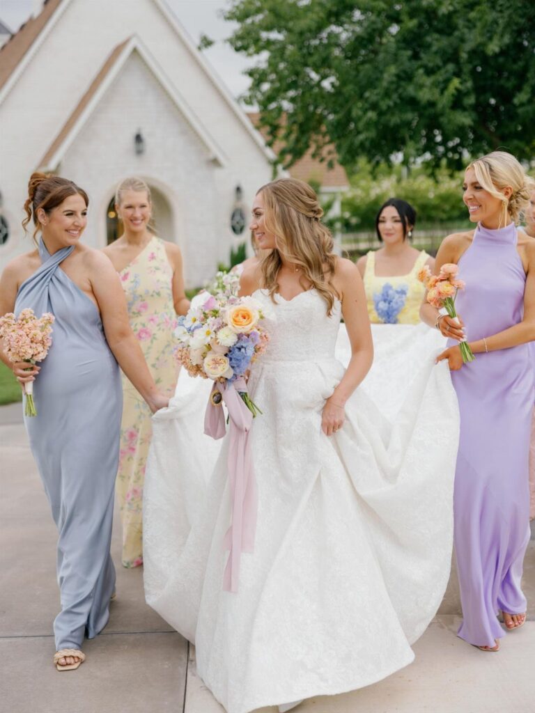 Bride in white gown walking with bridesmaids wearing blue, yellow, and lilac dresses.