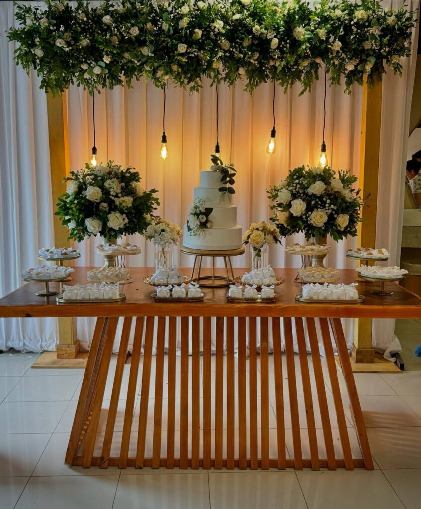Tiered white wedding cake on a wooden table with hanging warm light bulbs.