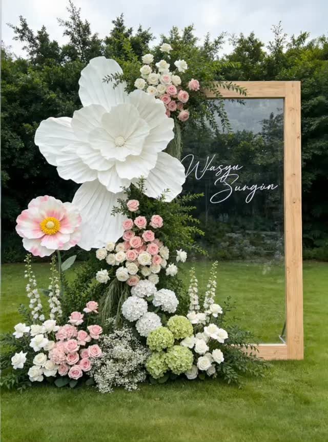 Wooden mirror frame decorated with giant white paper flowers and pink floral arrangements.