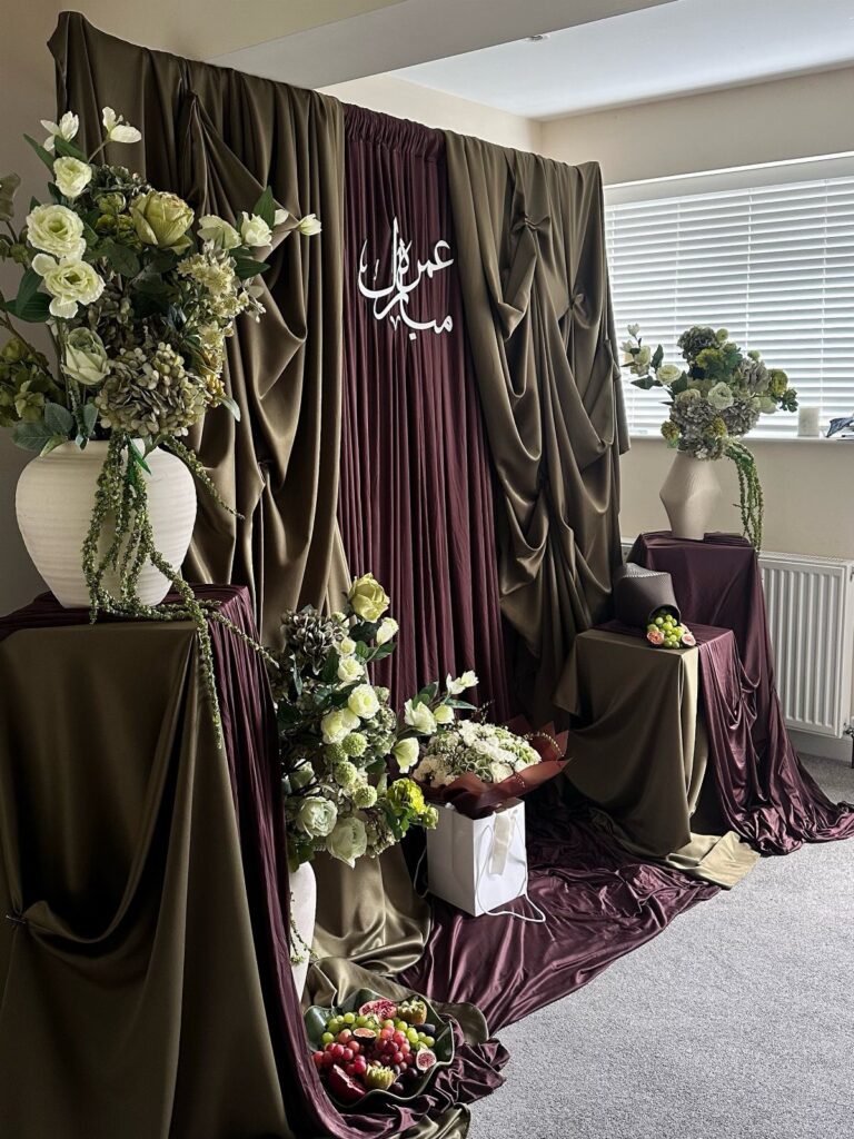 Olive green and burgundy draped fabric backdrop with white floral arrangements and calligraphy.