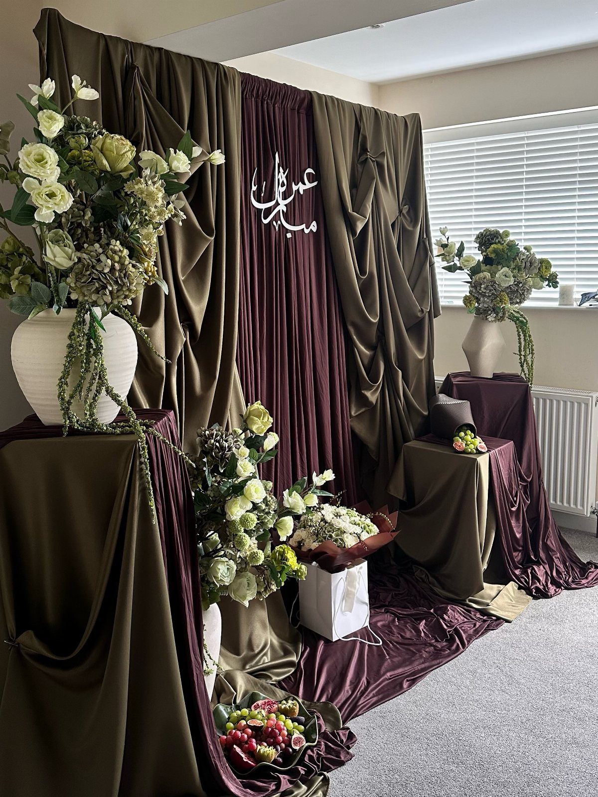 Olive green and burgundy draped fabric backdrop with white floral arrangements and calligraphy.