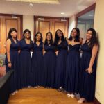 Seven women wearing long navy blue bridesmaid dresses posing together in a room.