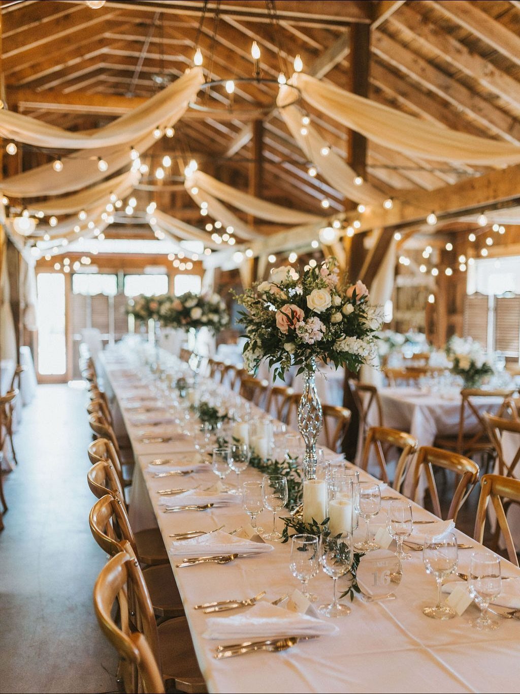 Long wedding table with white linens and neutral floral arrangements in a rustic barn.