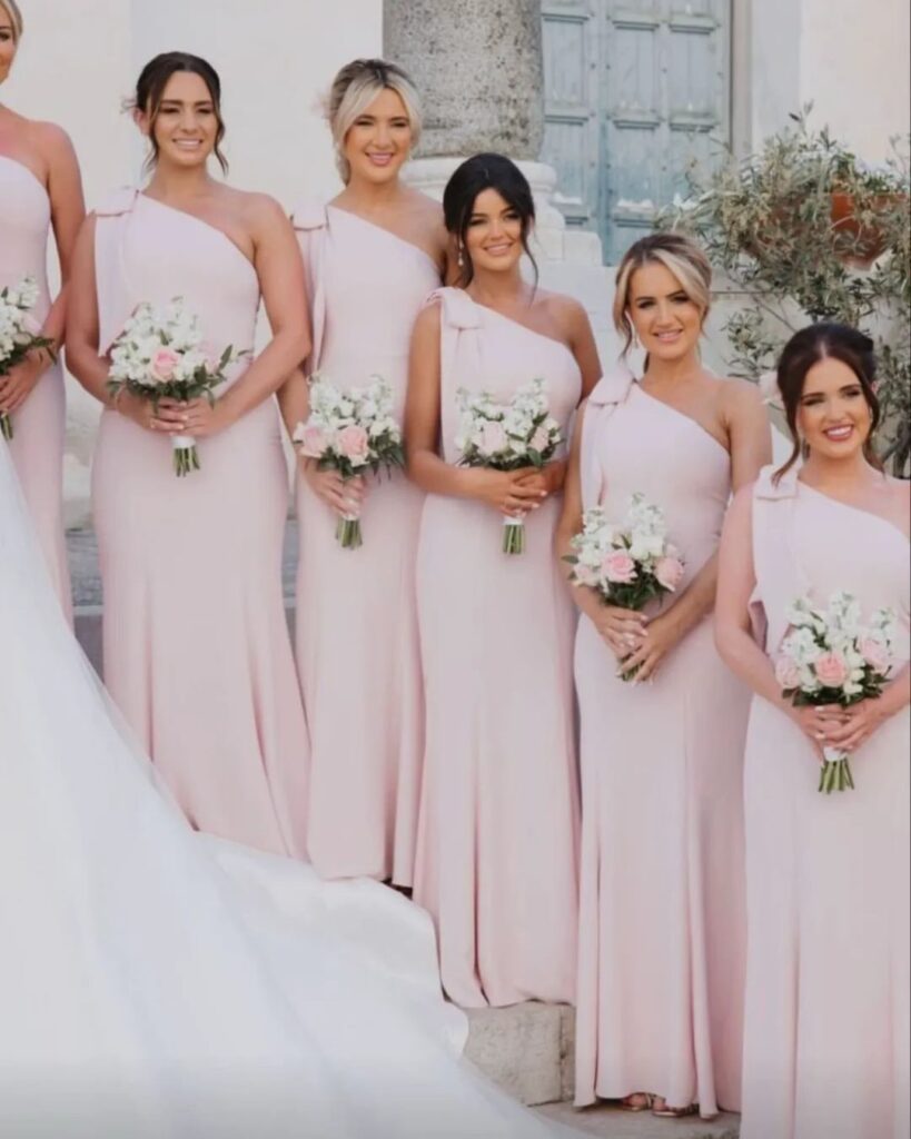 Bridesmaids in light pink one-shoulder dresses holding white and pink floral bouquets.