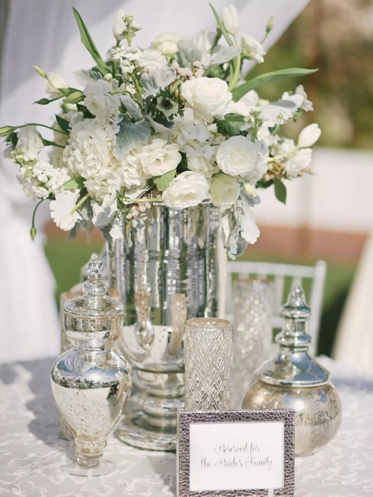 White floral centerpiece in a silver vase with silver decorative accents on a table.