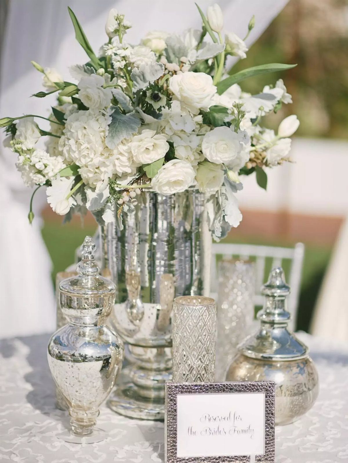 White floral centerpiece in a silver vase with silver decorative accents on a table.