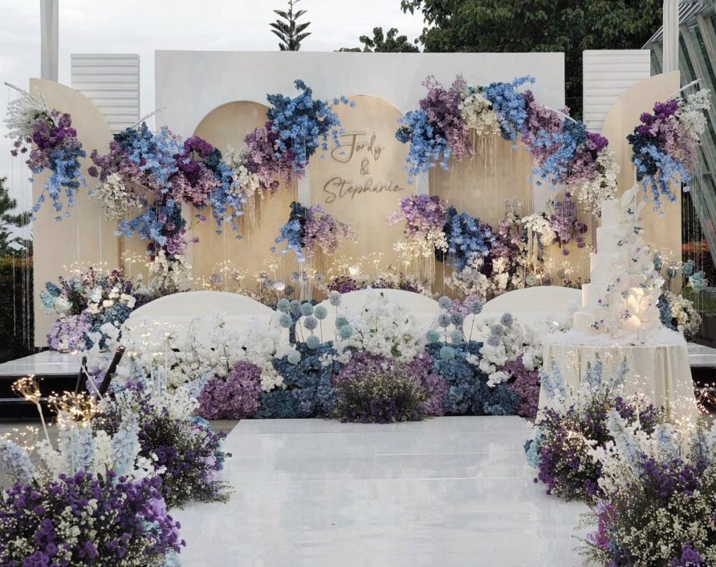 Wedding stage decorated with lush purple and blue flowers and a white backdrop.