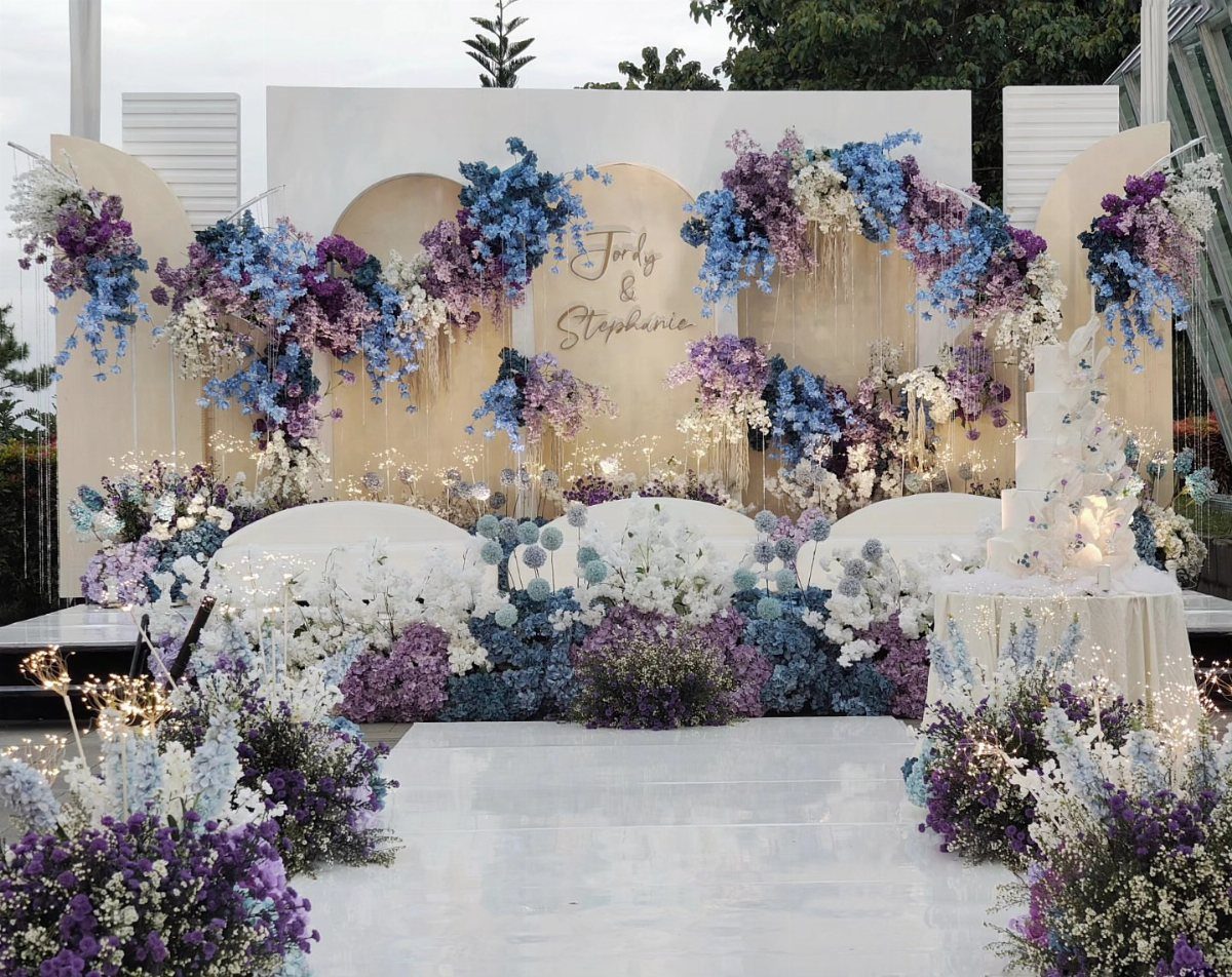 Wedding stage decorated with lush purple and blue flowers and a white backdrop.