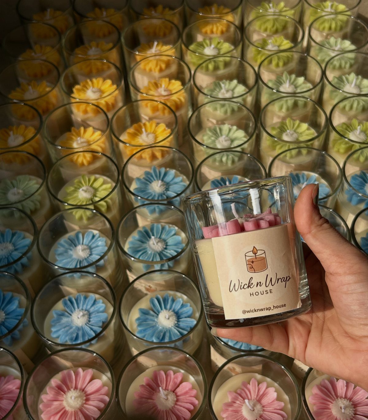 Hand holding a branded scented candle surrounded by colorful flower-shaped wax candles.
