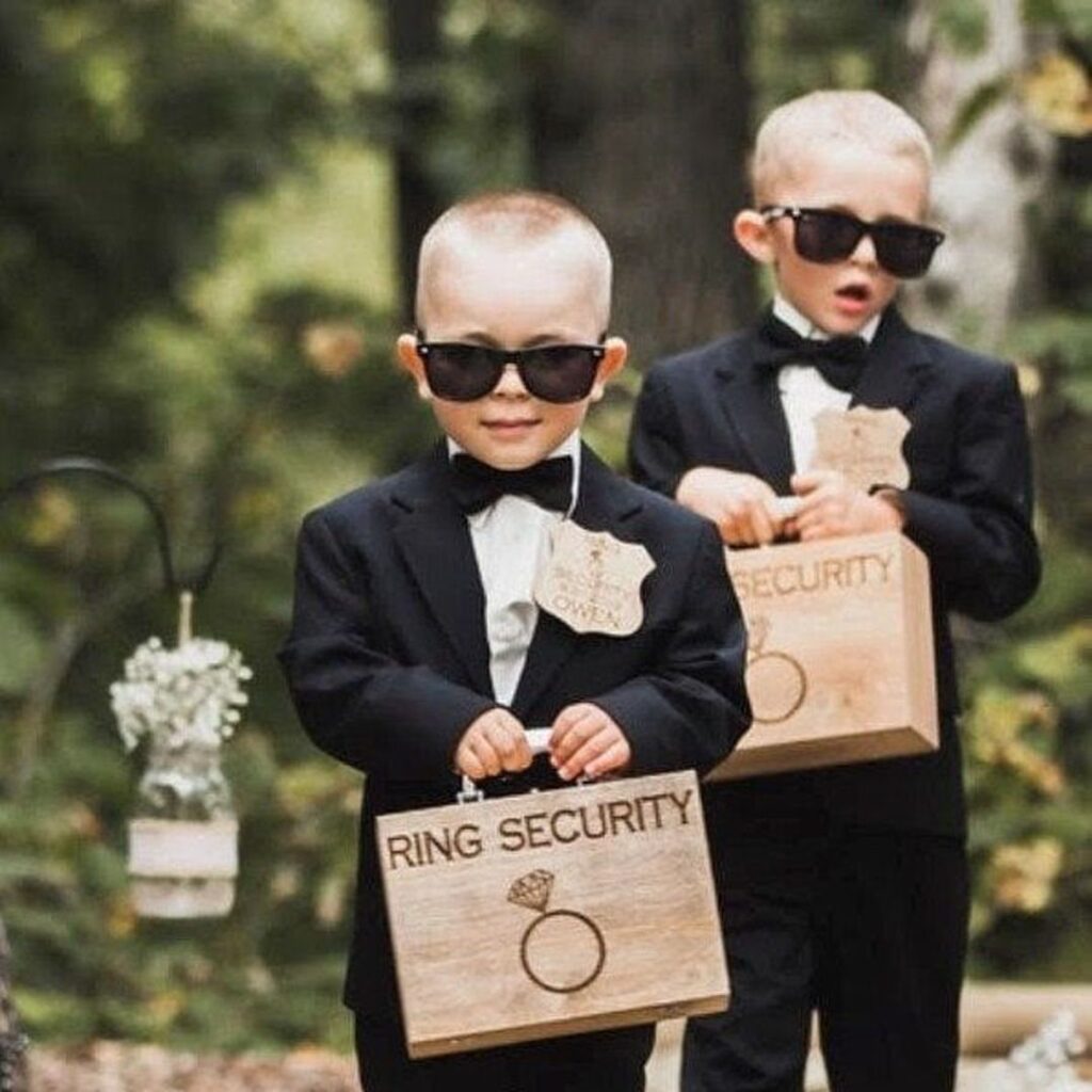 Two young ring bearers in tuxedos and sunglasses holding wooden ring security boxes.
