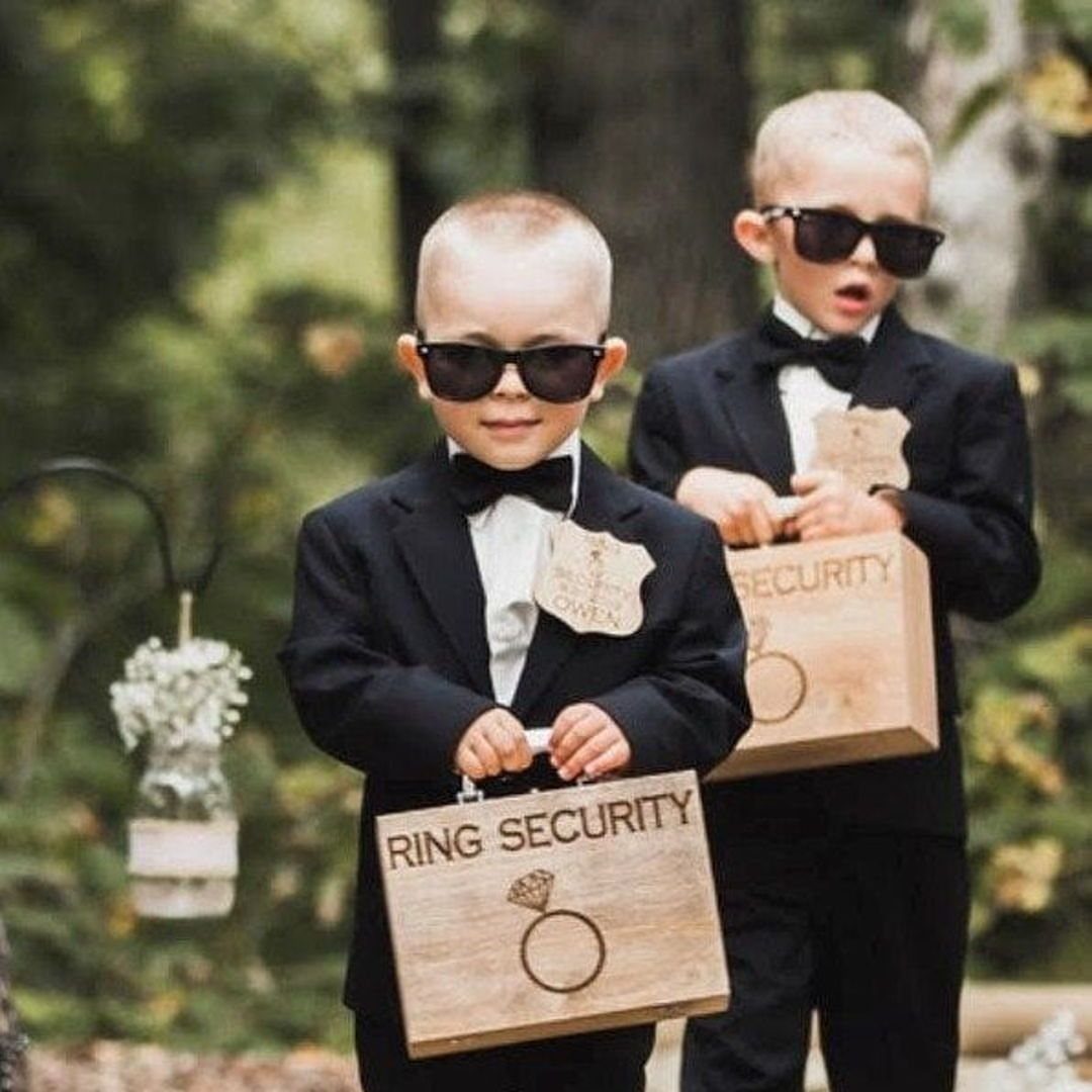 Two young ring bearers in tuxedos and sunglasses holding wooden ring security boxes.