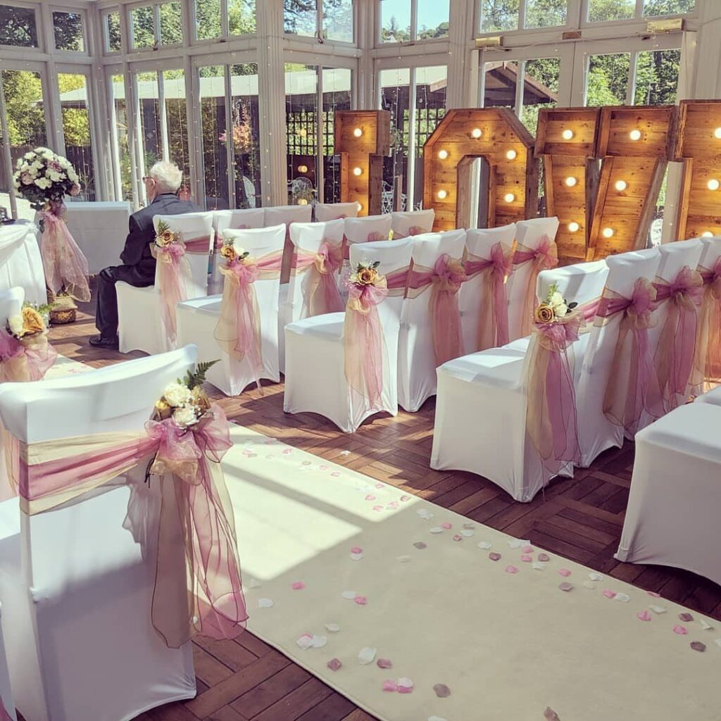 White wedding chairs with rose gold sheer sashes and floral accents in a sunlit room.