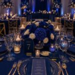 Royal blue wedding table setting with blue flowers, gold cutlery, and glowing candles.