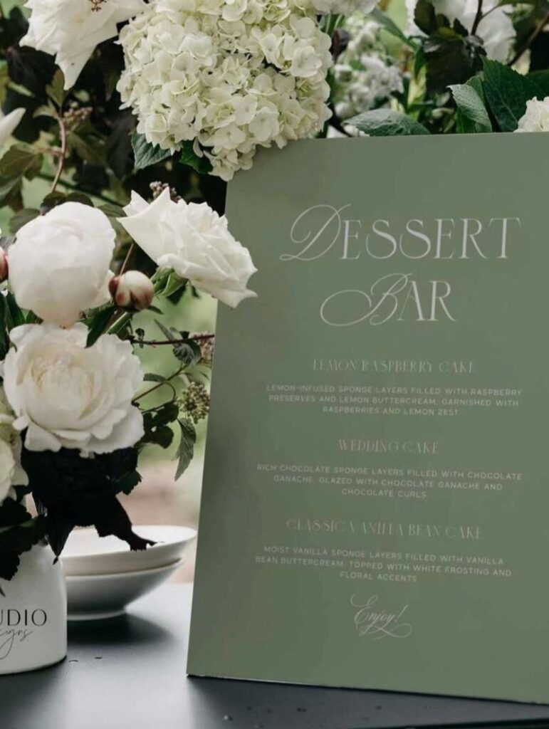 Sage green dessert bar menu card next to white roses and hydrangeas.