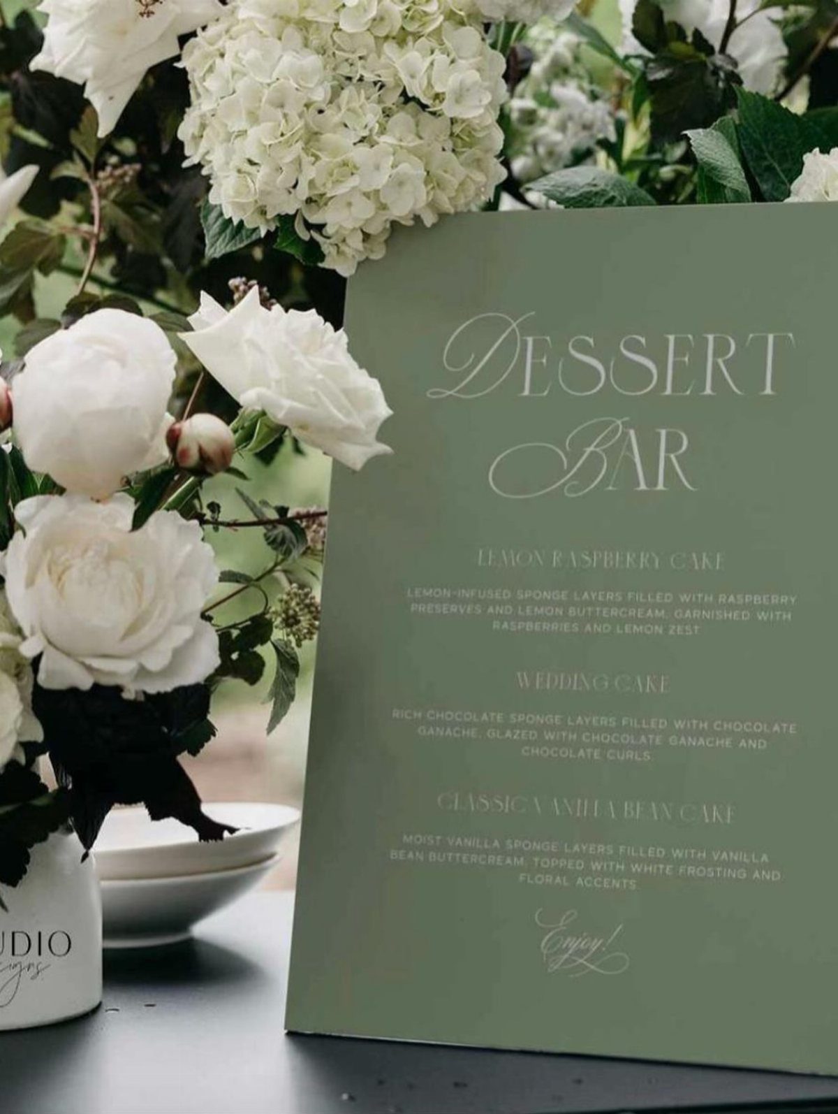 Sage green dessert bar menu card next to white roses and hydrangeas.