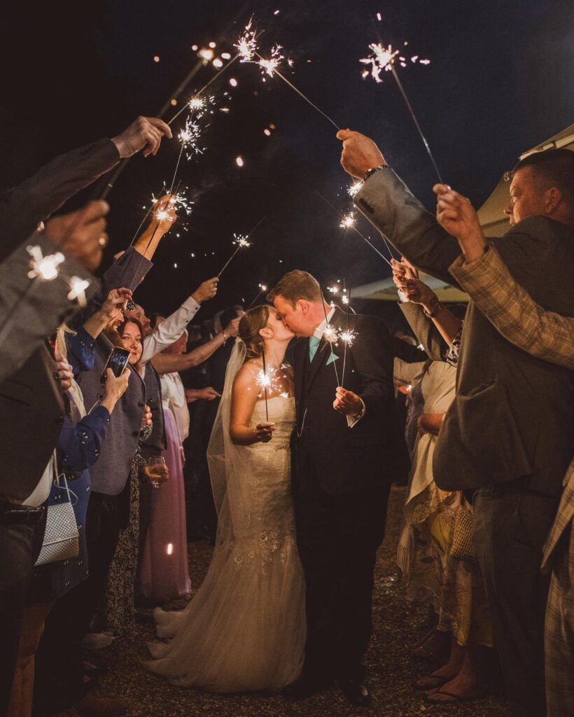 Bride and groom kissing while guests hold lit sparklers during a wedding send-off.