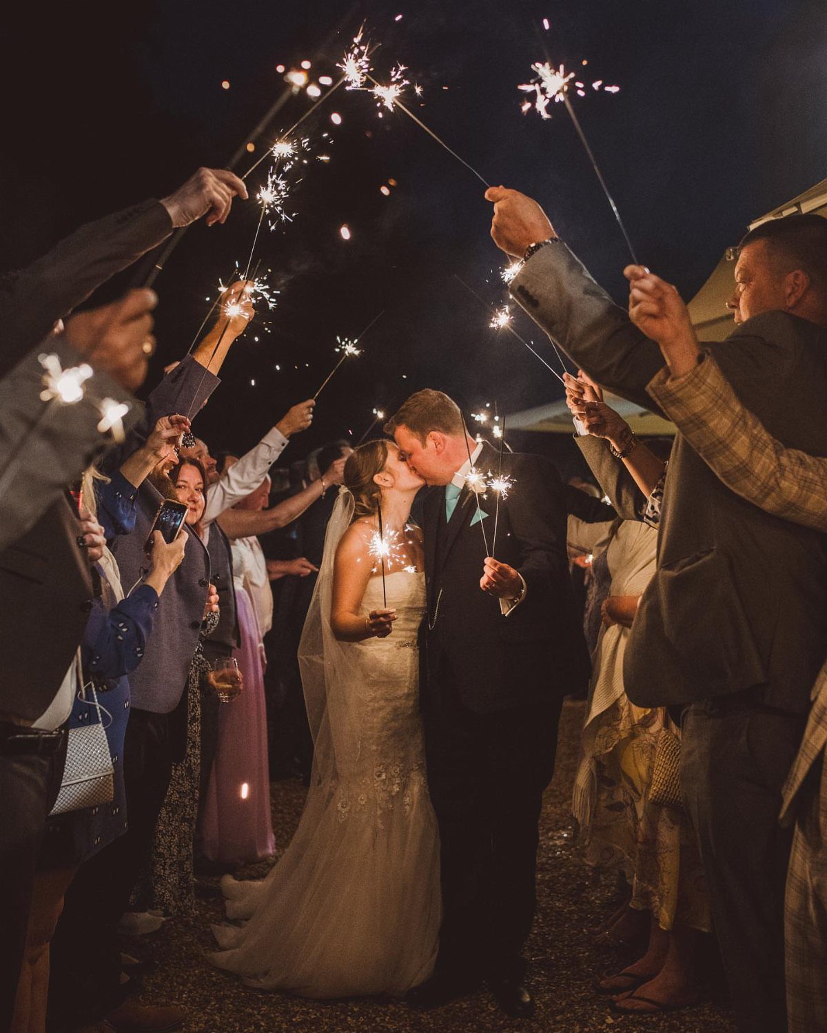 Bride and groom kissing while guests hold lit sparklers during a wedding send-off.