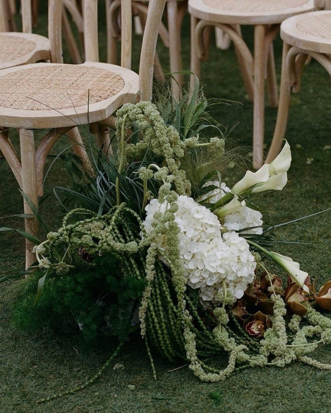 White and green floral arrangement on grass beside wooden wedding ceremony chairs.