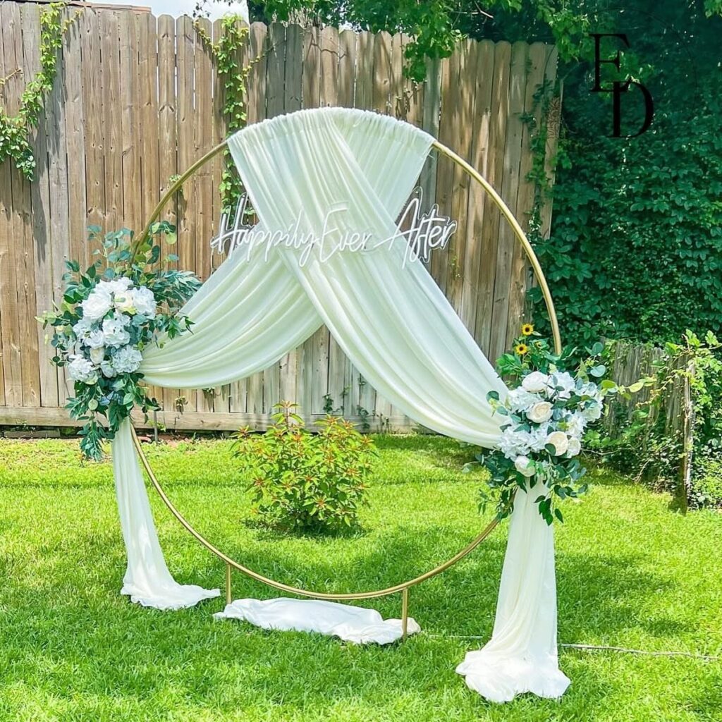 Gold circular wedding arch with white fabric draping and white floral arrangements outdoors