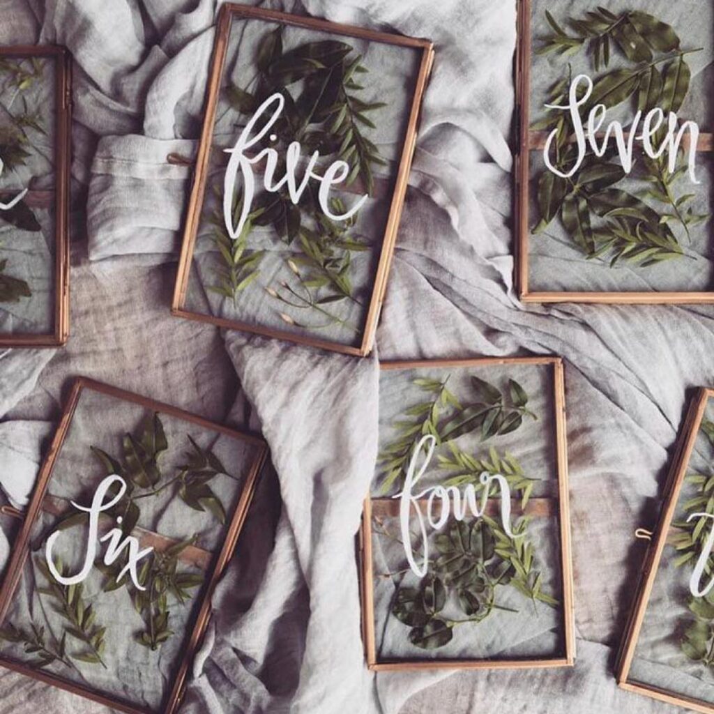 Glass table number frames with green leaves and white cursive text on grey fabric